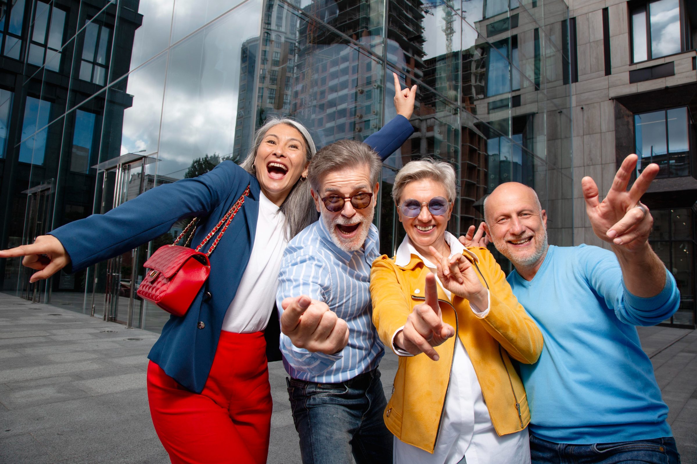 Four older friends laughing and posing playfully outside a glass city building