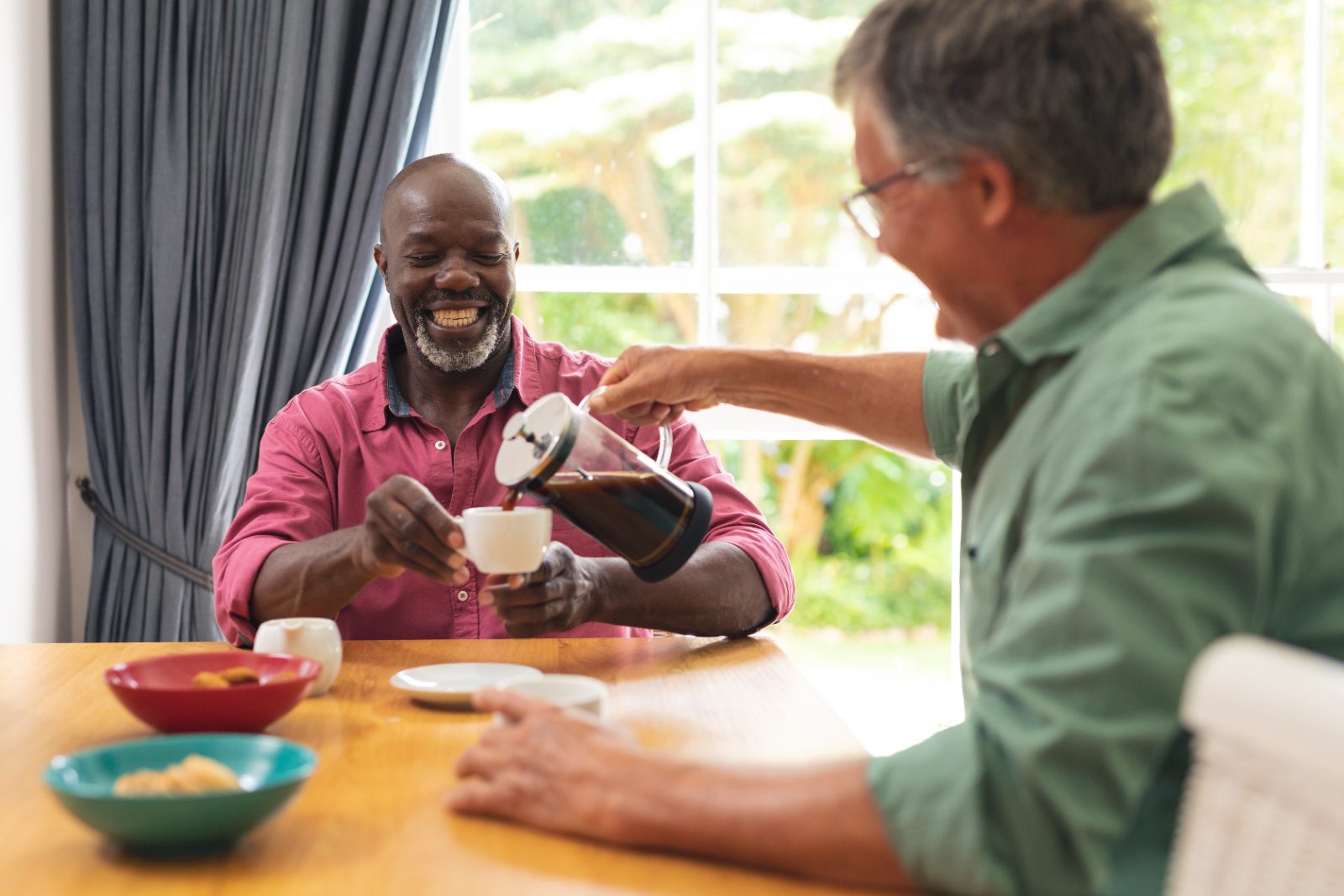 Two older friends at a kitchen table, one pouring coffee for the other, both smiling