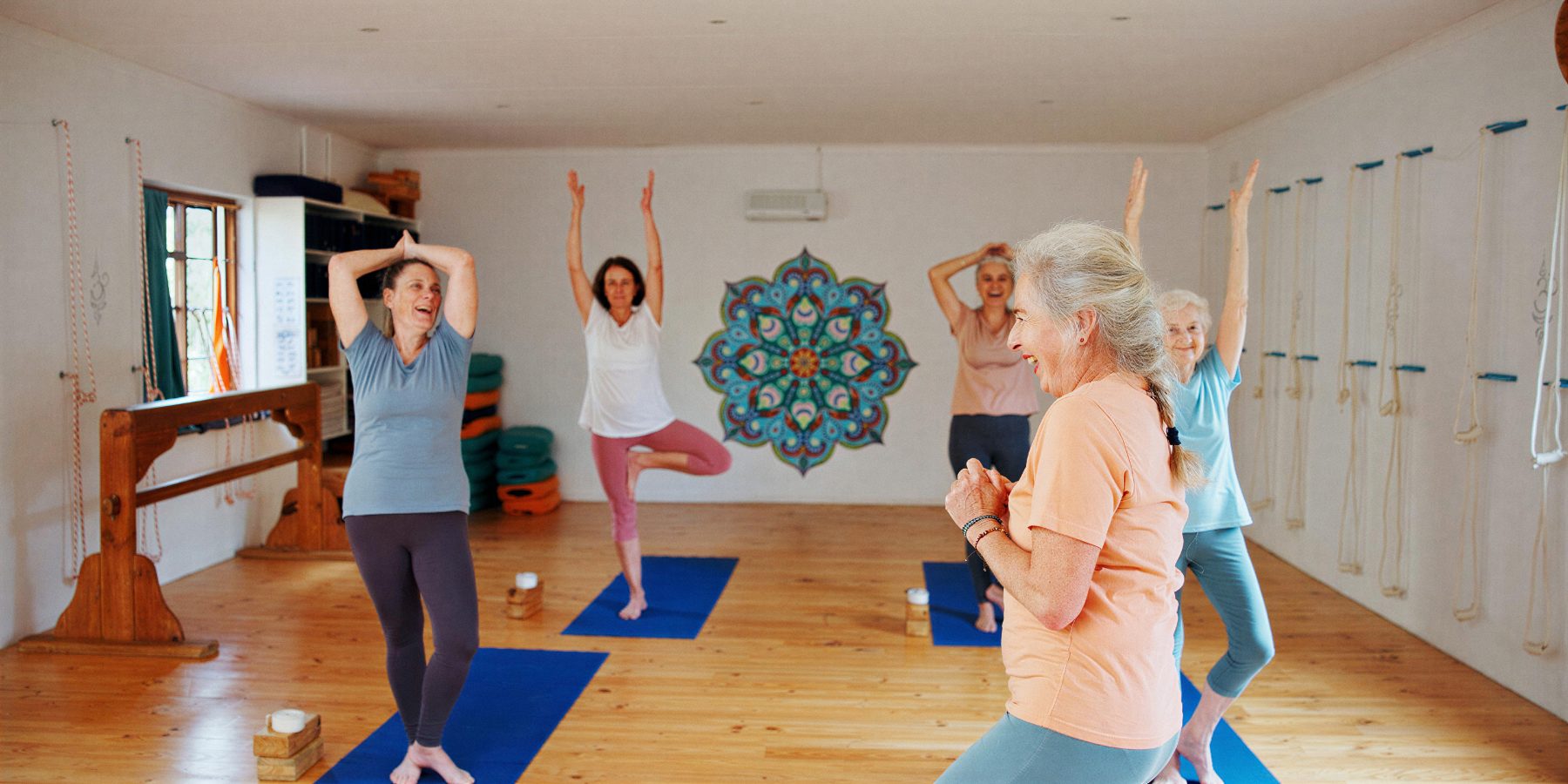 A smiling older woman in a peach top leads a small yoga class of women in a bright studio, all with arms raised and laughing